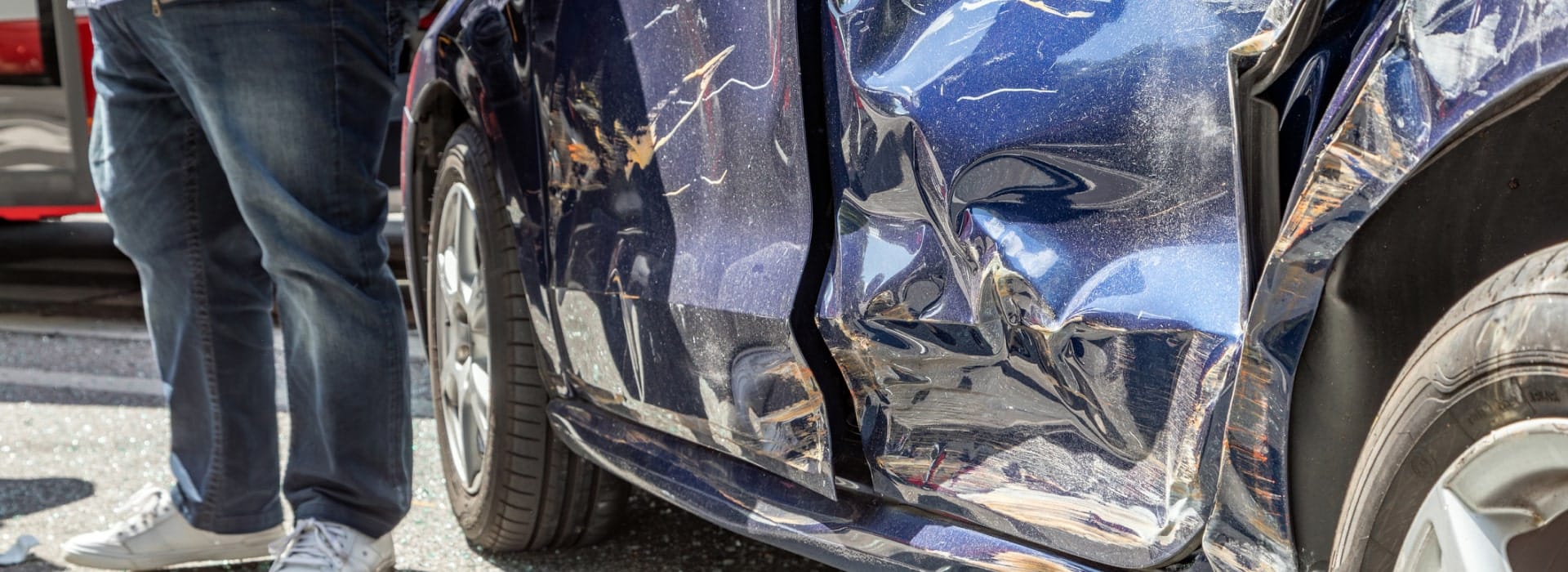 Caved-in driver's side door on a blue sedan, broken glass across the road